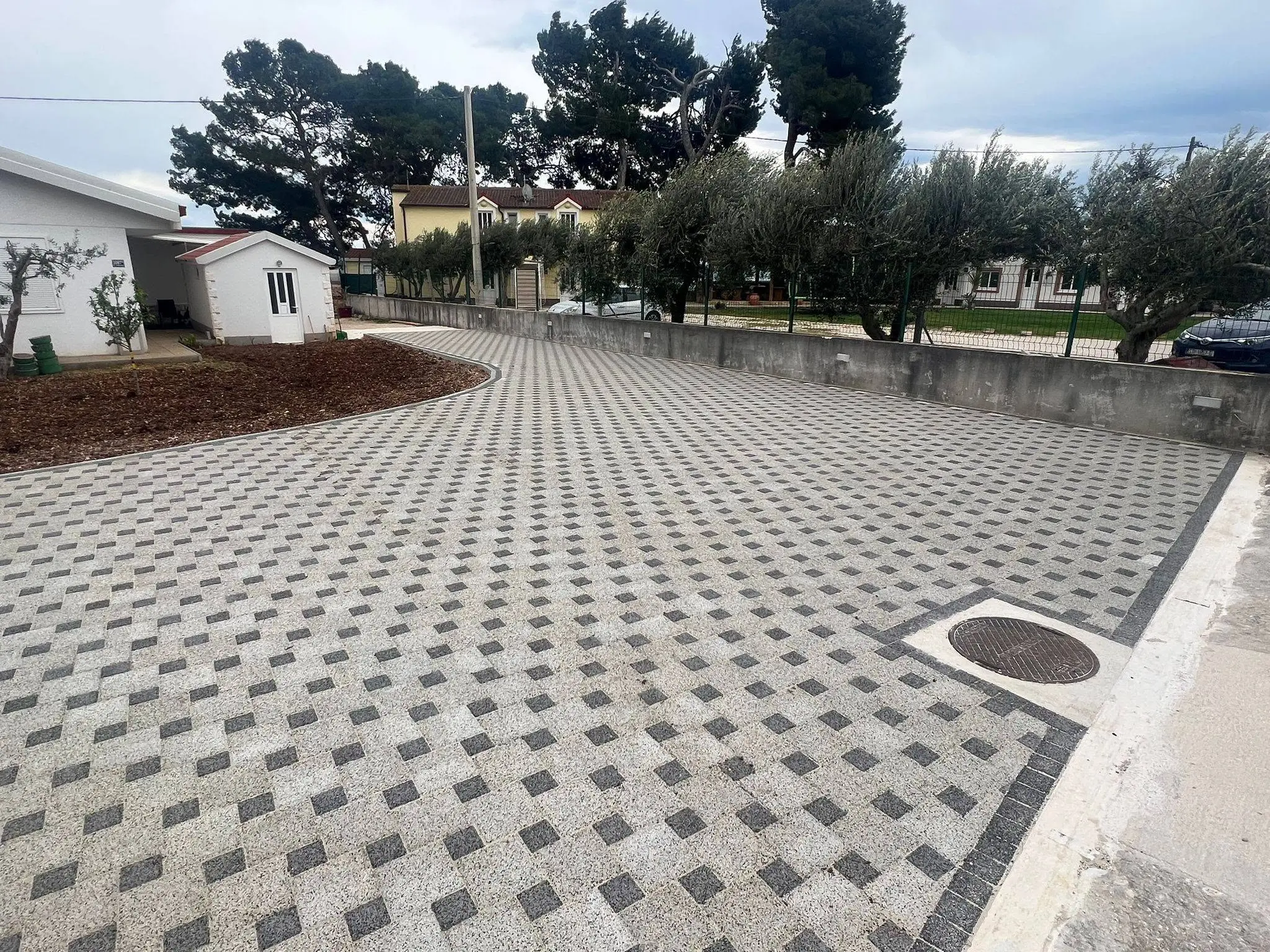 Prostrana površina popločana svijetlim opločnikom s tamnim kockama u uzorku, uz zid i red maslina.